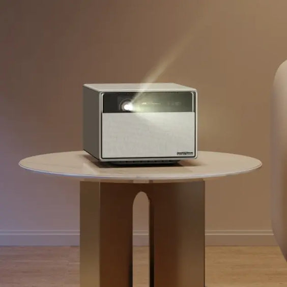 A modern, cube-shaped smart projector sitting on a round side table, emitting a beam of light.