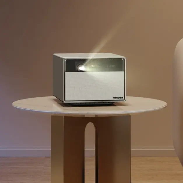 A modern, cube-shaped smart projector sitting on a round side table, emitting a beam of light.