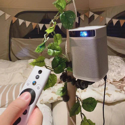 A small silver portable projector attached to a tent pole with decorative leaves, with a hand holding a remote control in the foreground, creating a cozy glamping atmosphere.